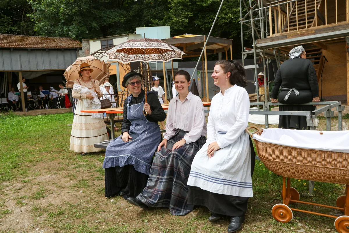 Dass sich die Fabrikantenfrau Louisa Maggi (Barbara Hügi, links hinten mit Baby im Arm) unter die Arbeiterinnen (Angela Bürki, Céline Jacquat und Leonie Mauchle) mischt, sorgt bei diesen für Überraschung. Man sieht drei Frauen in alten Kostümen auf einer Bank sitzen.